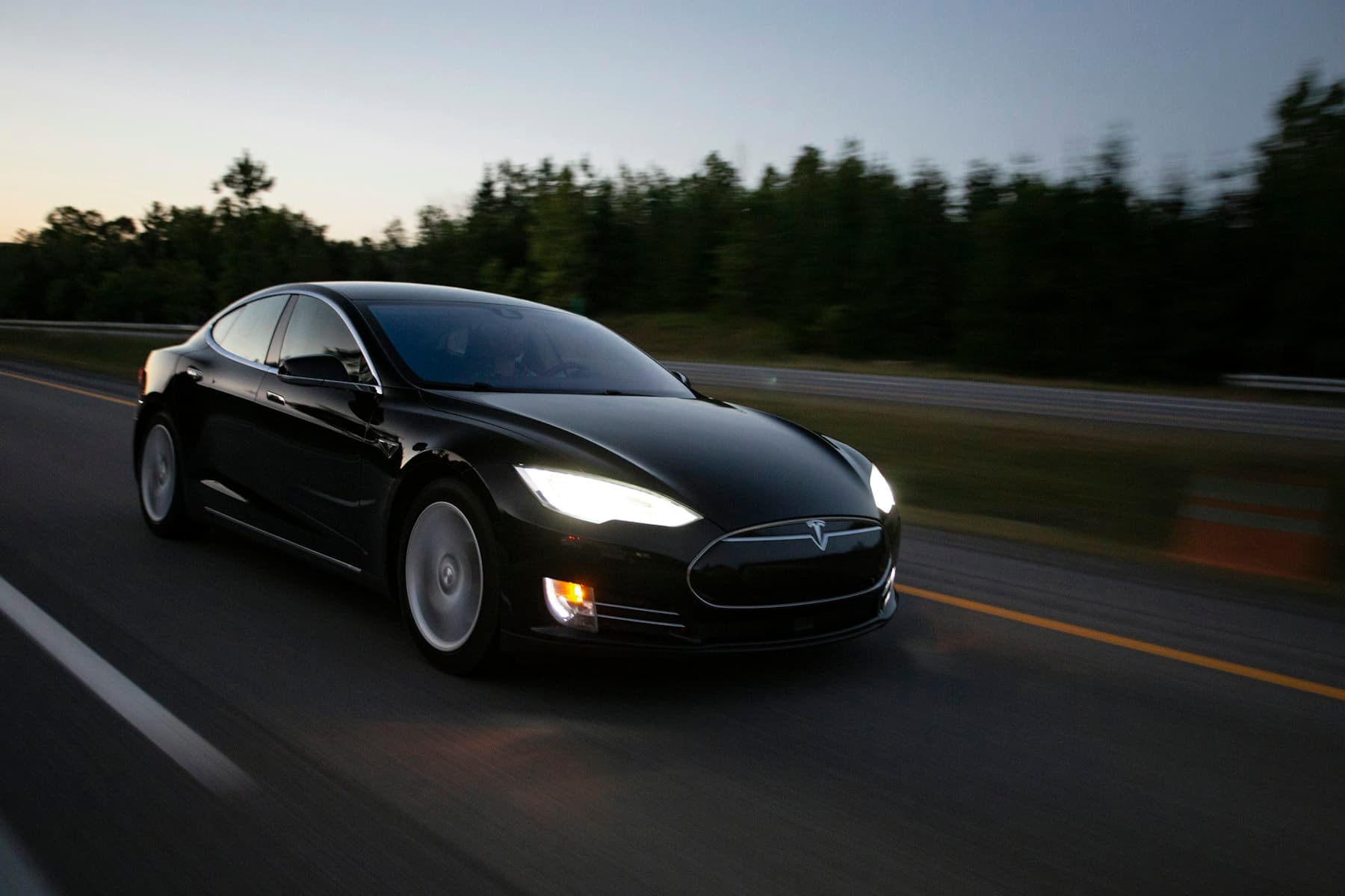Black Tesla Model S driving along a tree-lined road with headlights on