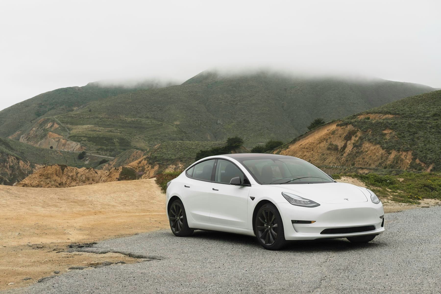 White Tesla Model 3 parked on a gravel verge with misty green hills behind